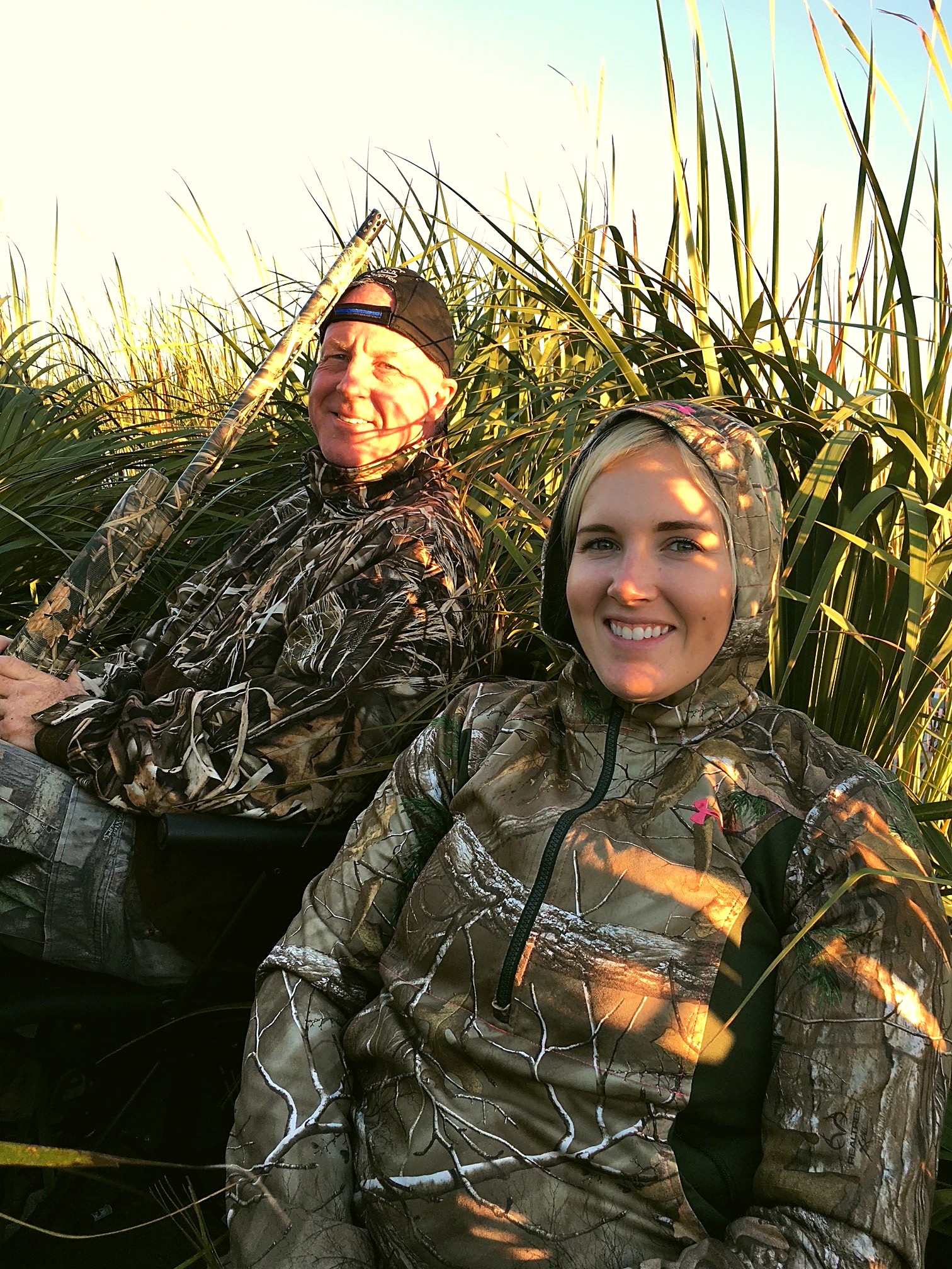 Two people sit together in a duck blind among tall marsh grass.
